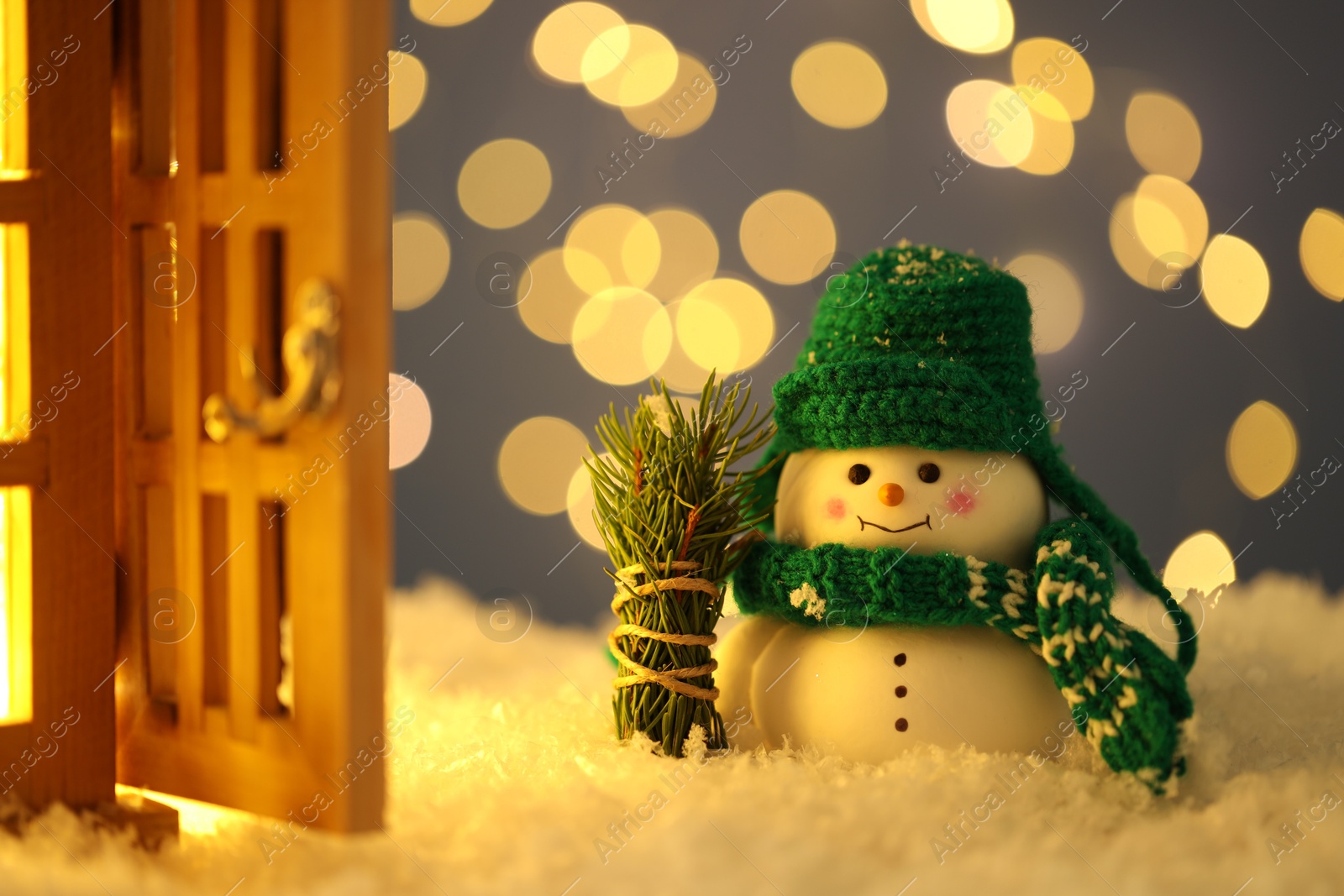 Photo of Cute decorative snowman, fir tree branches and lantern on artificial snow against blurred lights, closeup