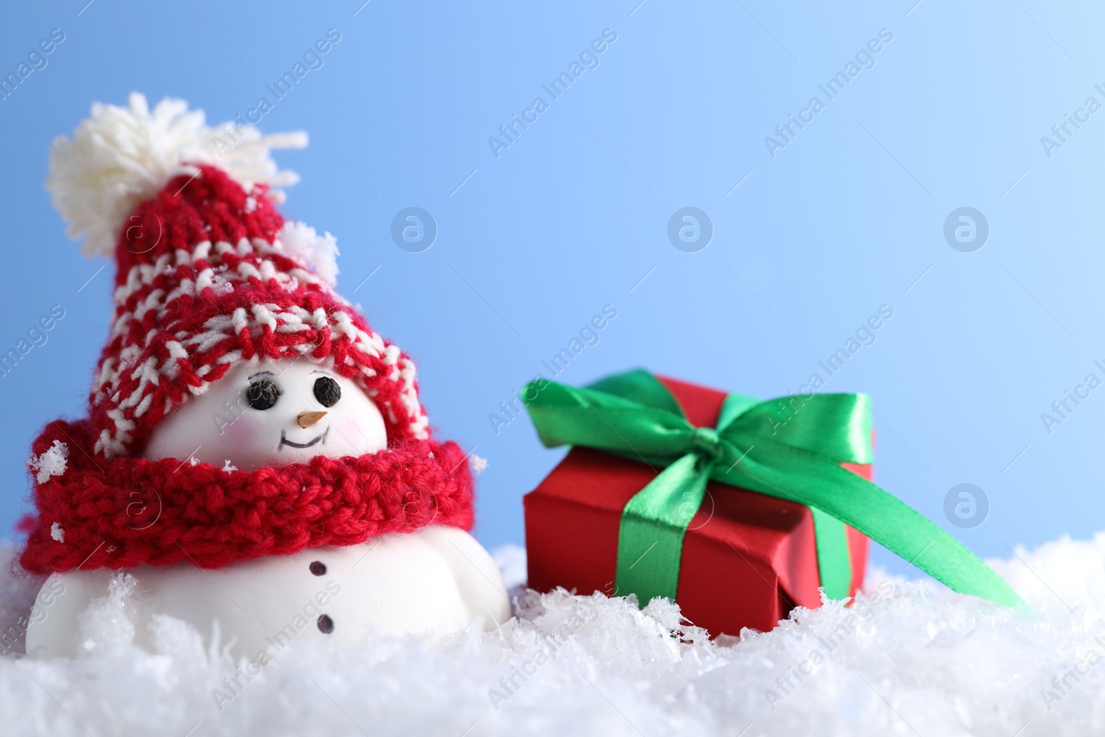 Photo of Cute decorative snowman and gift box on artificial snow against light blue background, closeup