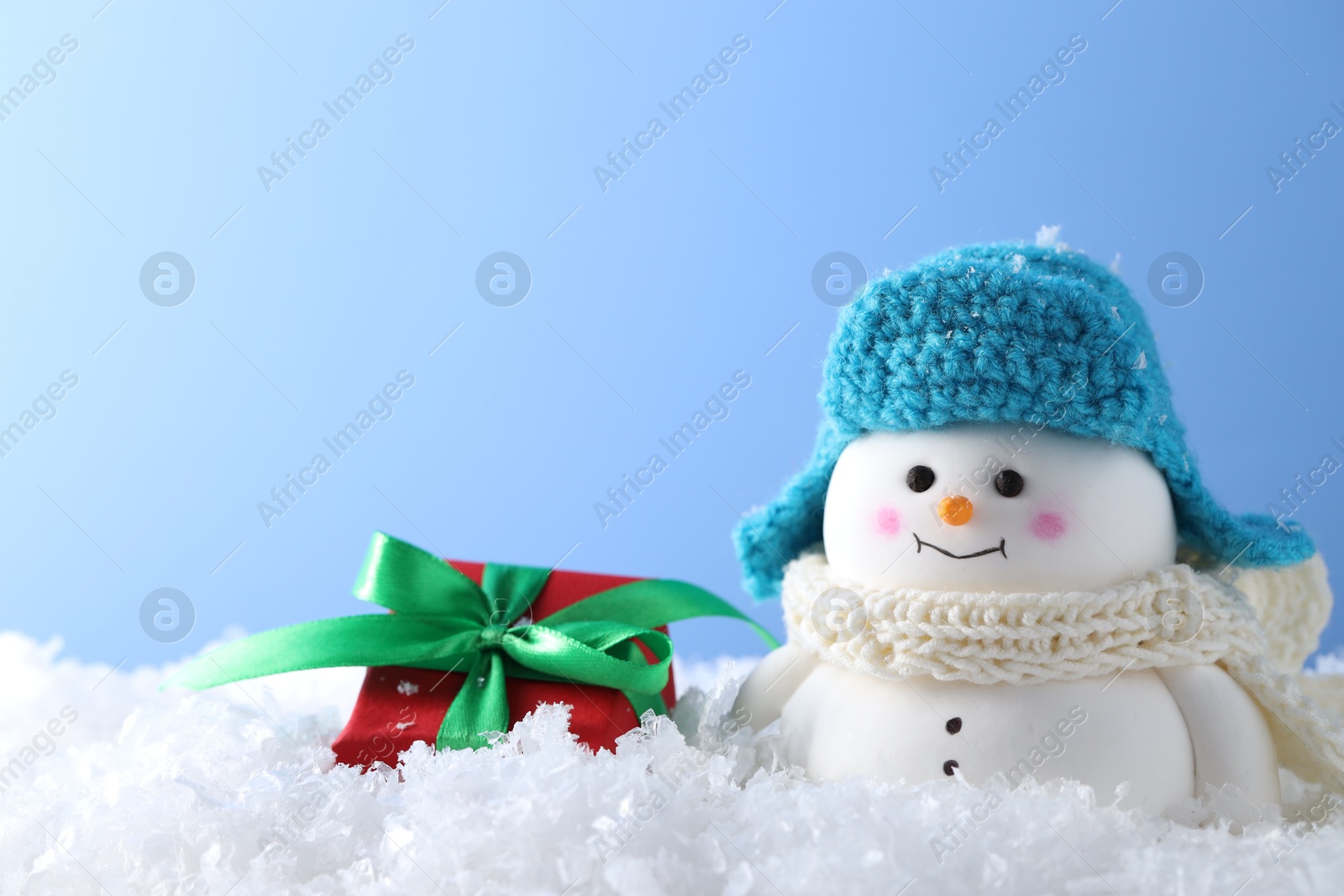 Photo of Cute decorative snowman and gift box on artificial snow against light blue background, closeup. Space for text