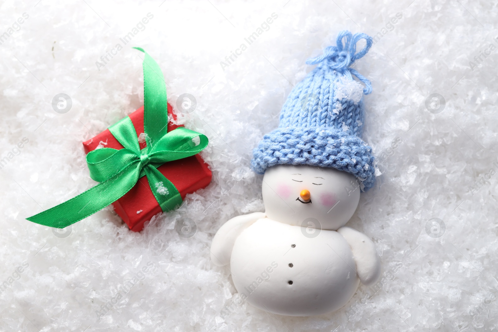 Photo of Cute decorative snowman and gift box on artificial snow, flat lay