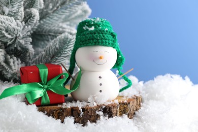 Photo of Cute decorative snowman, gift box and fir tree branches on artificial snow against light blue background