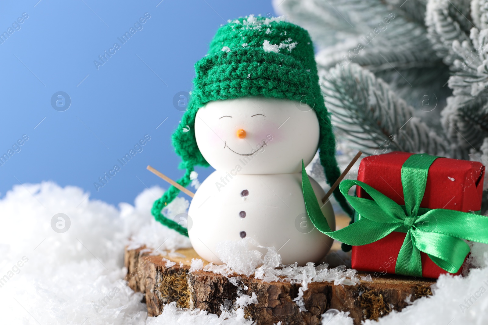 Photo of Cute decorative snowman, gift box and fir tree branches on artificial snow against light blue background, closeup