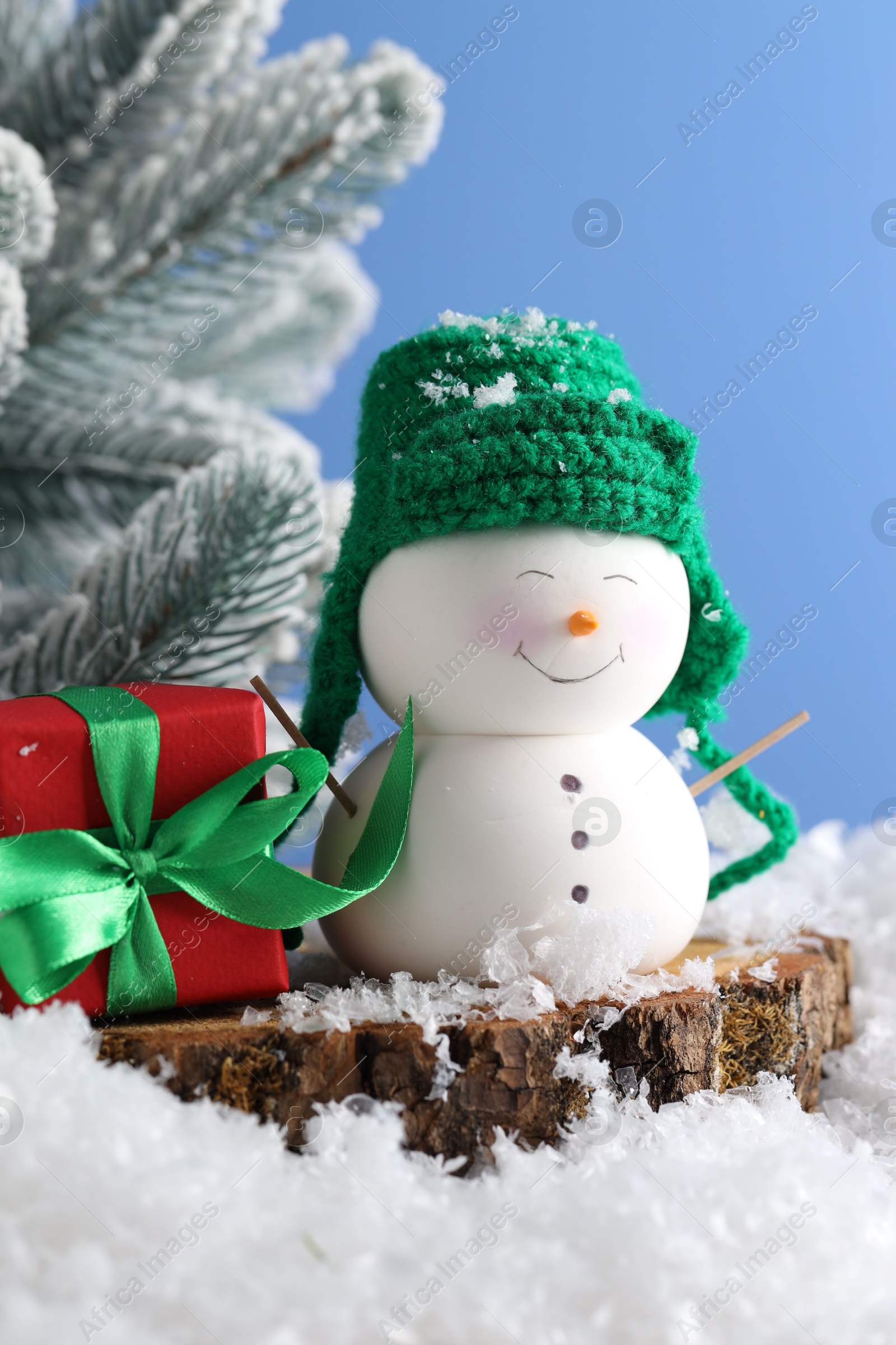 Photo of Cute decorative snowman, gift box and fir tree branches on artificial snow against light blue background, closeup