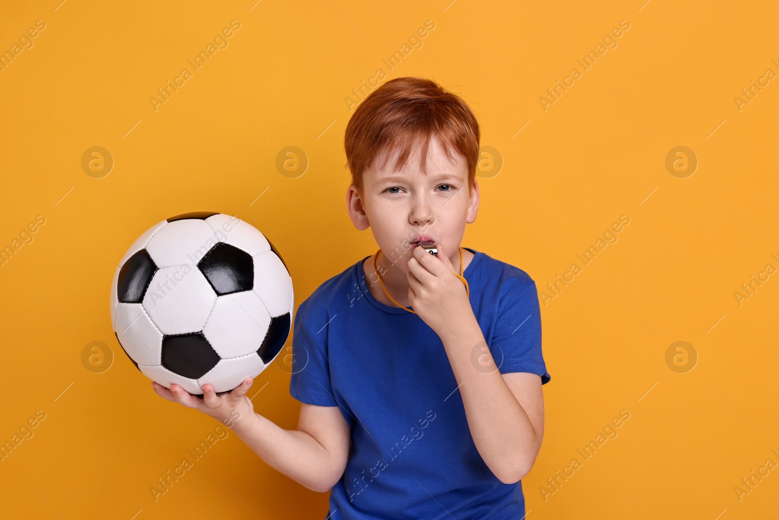 Photo of Little boy with soccer ball blowing whistle on orange background