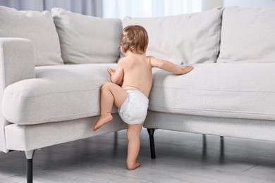 Little child in diaper climbing on sofa at home, back view