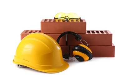 Photo of Yellow hard hat, red bricks, goggles and earmuffs isolated on white. Building material and safety equipment