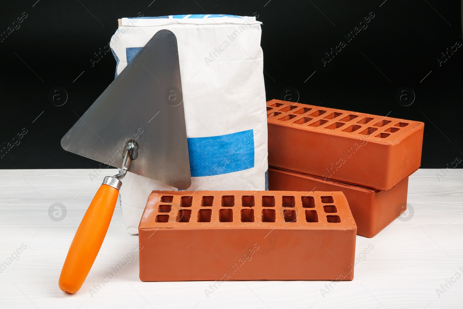 Photo of Cement powder in bag, trowel and red bricks on white wooden table. Building materials and construction tools