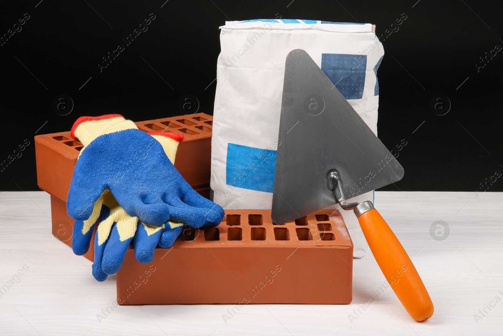 Photo of Cement powder in bag, gloves, trowel and red bricks on white wooden table. Building materials and construction tools