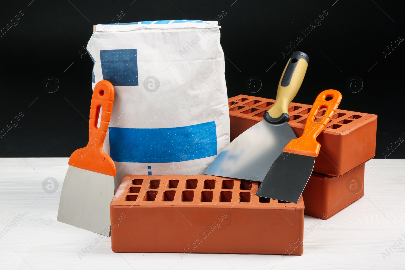 Photo of Cement powder in bag, putty knives and red bricks on white wooden table. Building materials and construction tools