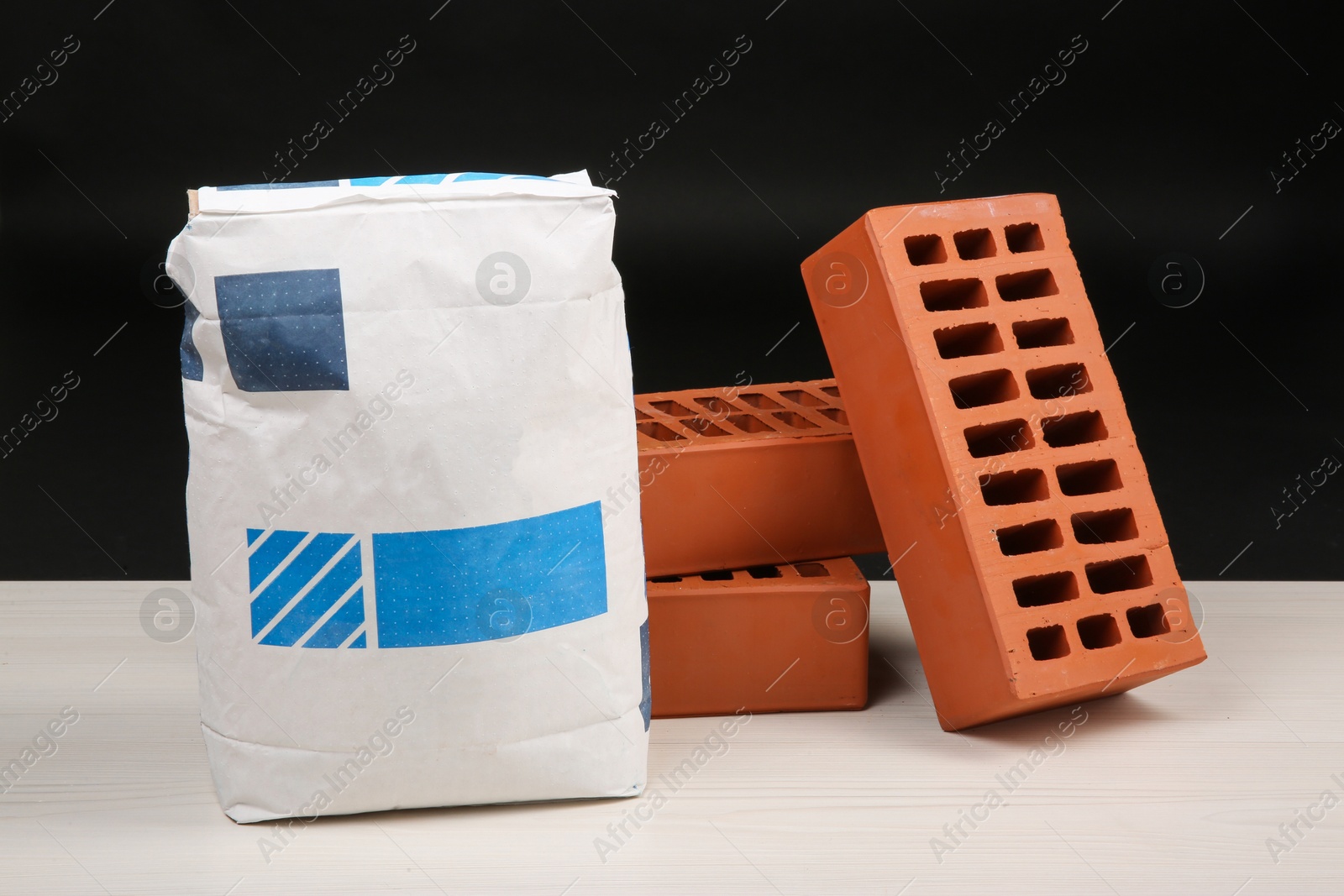 Photo of Red bricks and bag of cement on light wooden table against black background. Building materials