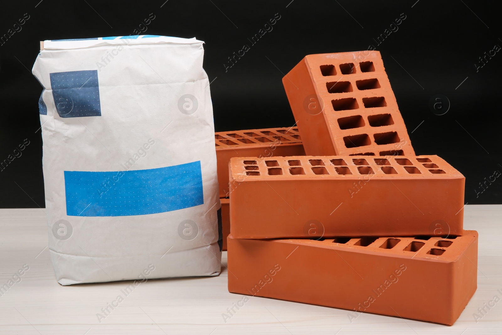 Photo of Red bricks and bag of cement on light wooden table against black background. Building materials