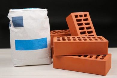 Red bricks and bag of cement on light wooden table against black background. Building materials