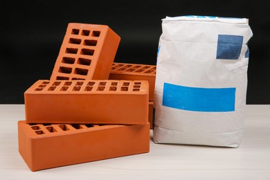 Photo of Red bricks and bag of cement on light wooden table against black background. Building materials