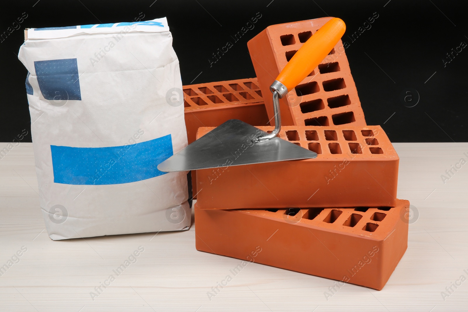 Photo of Red bricks, bag of cement and putty knife on light wooden table against black background. Building materials