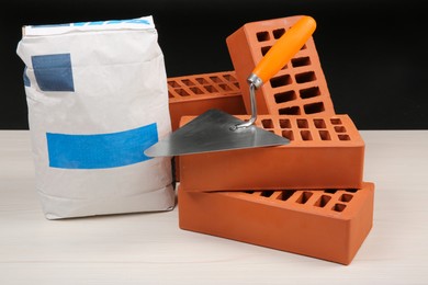 Red bricks, bag of cement and putty knife on light wooden table against black background. Building materials