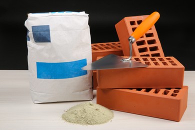 Red bricks, bag of cement and putty knife on light wooden table against black background. Building materials