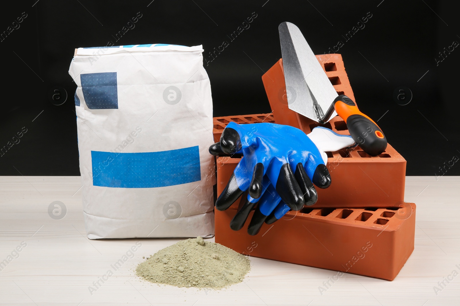 Photo of Red bricks, cement powder, putty knife and gloves on light wooden table against black background. Building materials