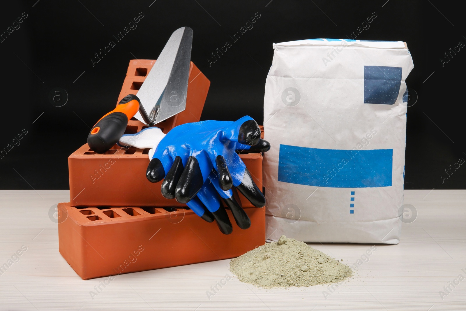 Photo of Red bricks, cement powder, putty knife and gloves on light wooden table against black background. Building materials