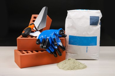 Photo of Red bricks, cement powder, putty knife and gloves on light wooden table against black background. Building materials