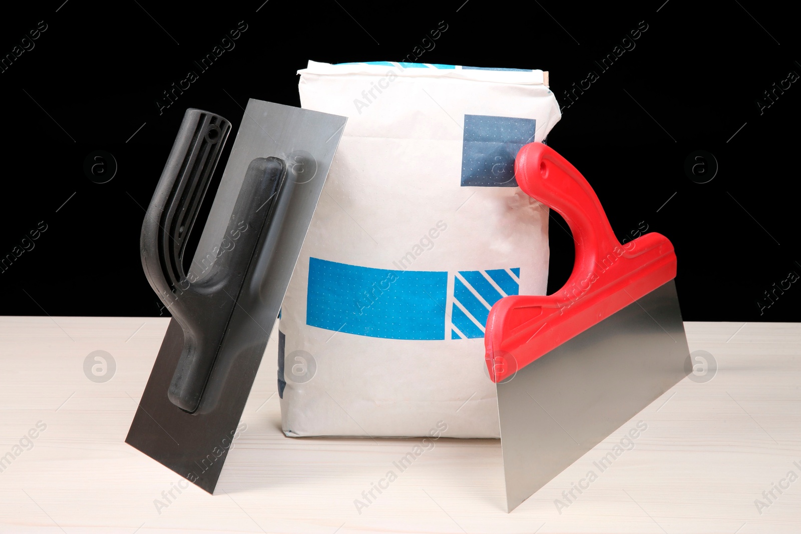 Photo of Bag of cement powder and putty knives on light wooden table against black background. Building material and construction tools