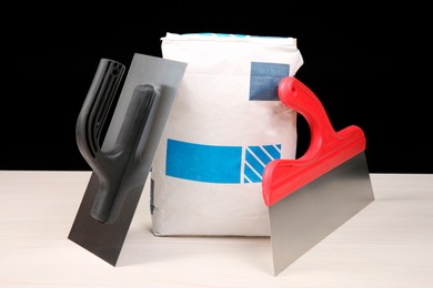 Photo of Bag of cement powder and putty knives on light wooden table against black background. Building material and construction tools