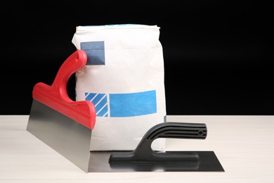 Photo of Bag of cement powder and putty knives on light wooden table against black background. Building material and construction tools