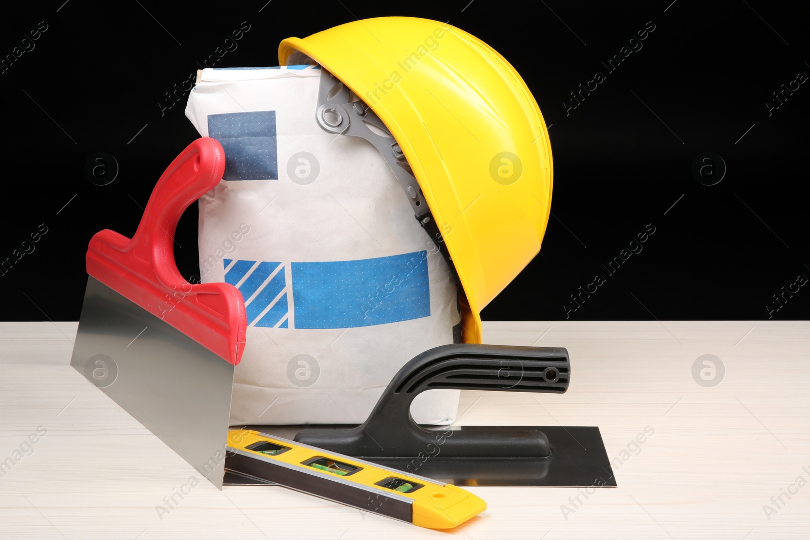 Photo of Yellow hard hat, bag of cement powder, putty knives and spirit level on light wooden table against black background. Building material and construction tools