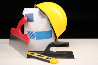 Yellow hard hat, bag of cement powder, putty knives and spirit level on light wooden table against black background. Building material and construction tools