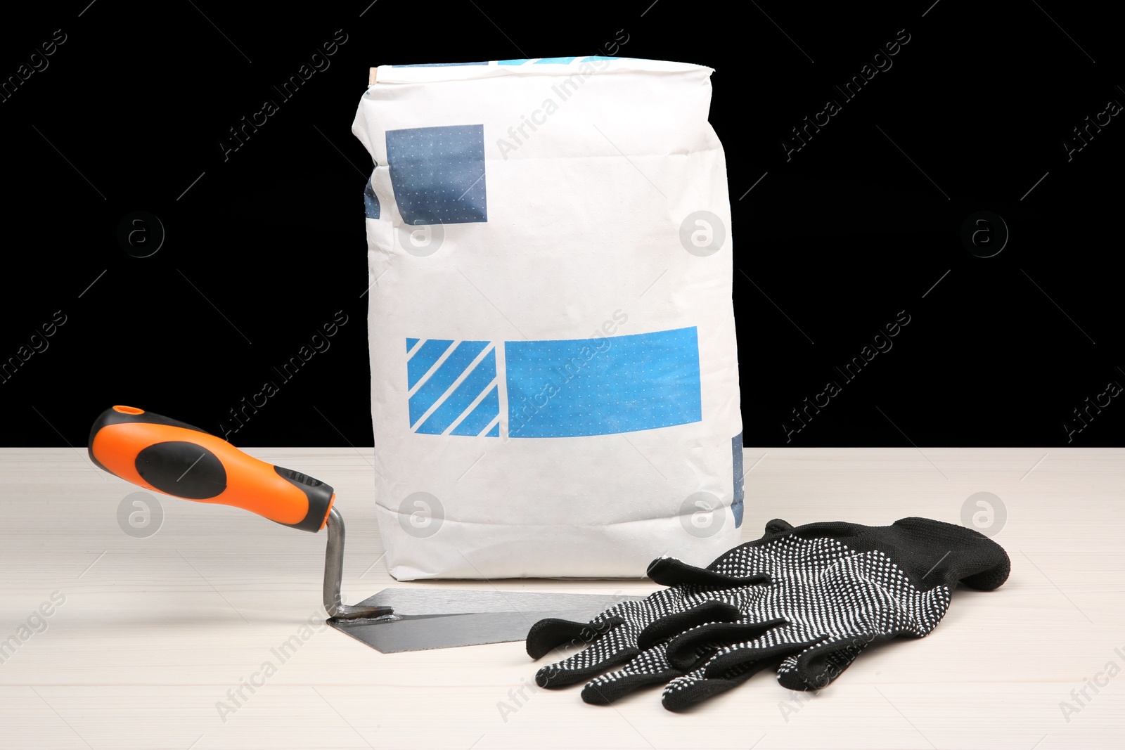 Photo of Bag of cement powder, putty knife and rubber gloves on light wooden table against black background. Building material