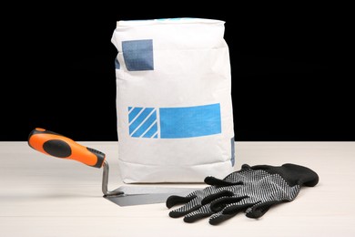 Bag of cement powder, putty knife and rubber gloves on light wooden table against black background. Building material