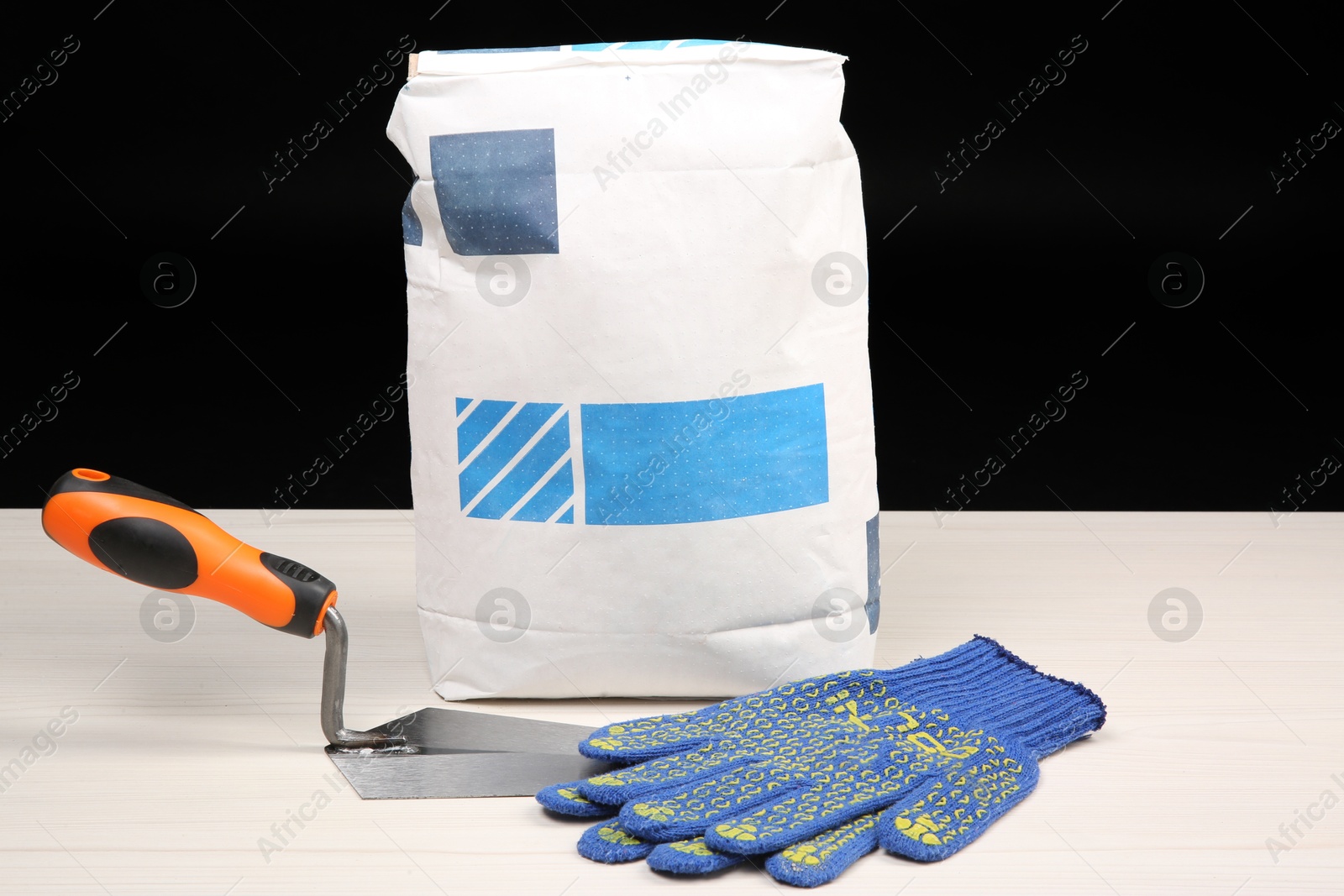 Photo of Bag of cement powder, putty knife and rubber gloves on light wooden table against black background. Building material