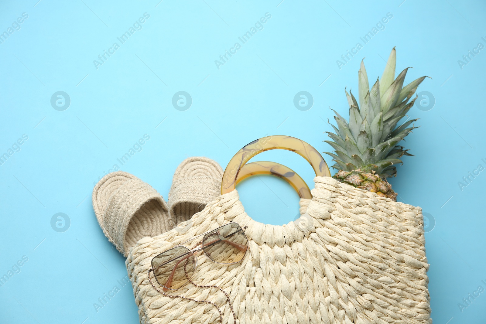 Photo of Elegant woman's straw bag with shoes, sunglasses and pineapple on light blue background, top view