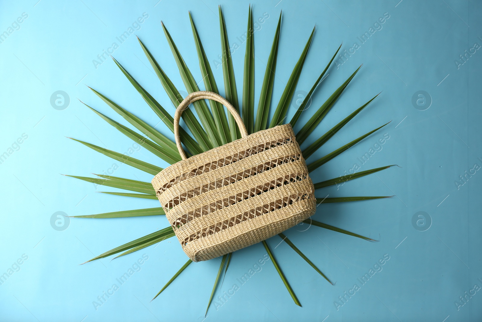 Photo of Elegant woman's straw bag with tropical leaf on light blue background, top view