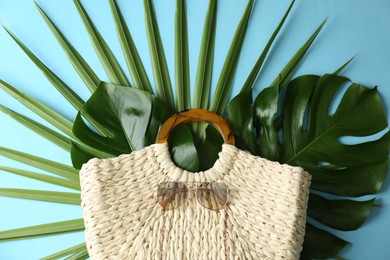 Photo of Elegant woman's straw bag with tropical leaves and sunglasses on light blue background, top view