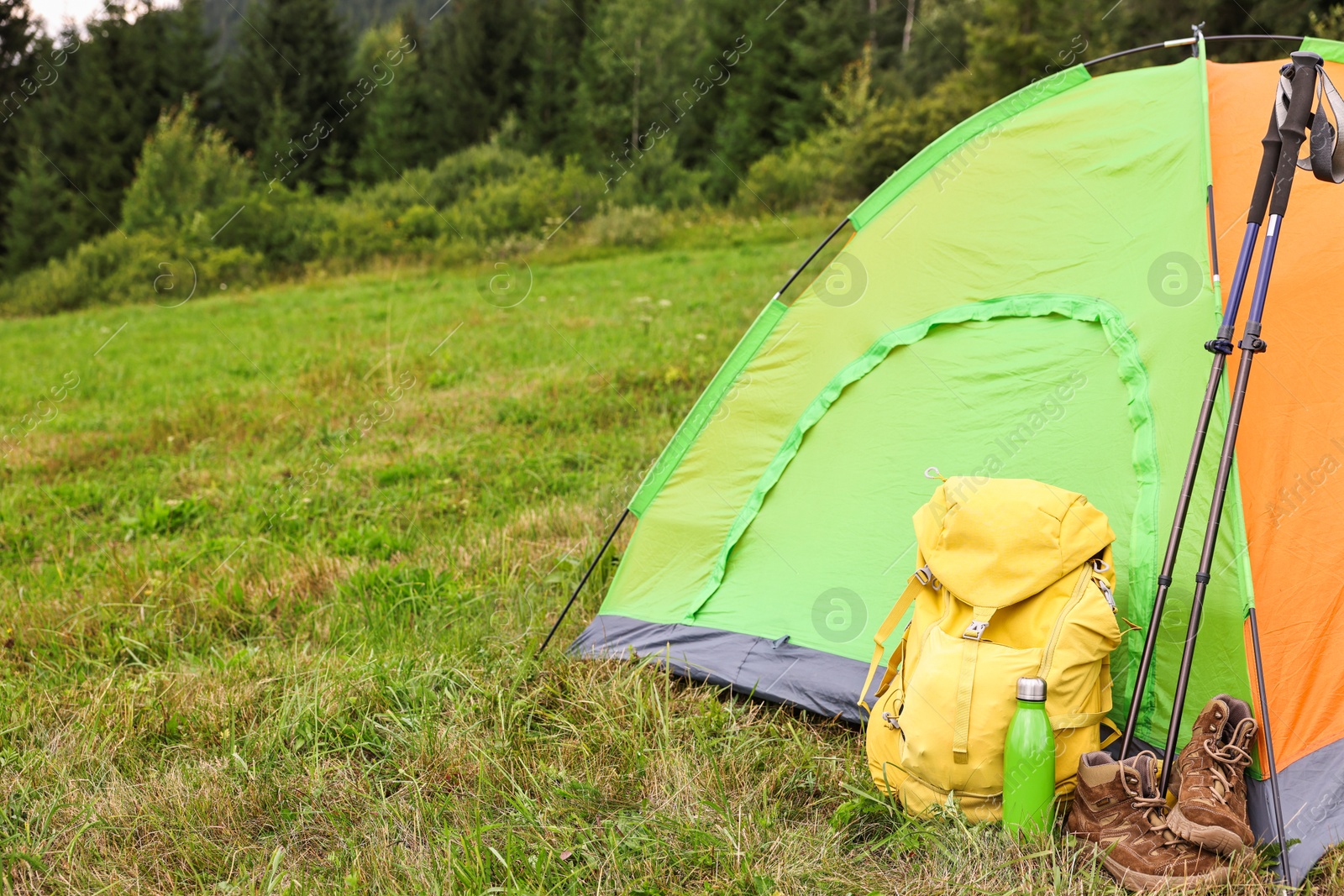 Photo of Tent, backpack, trekking poles and thermos on green grass in mountains, space for text