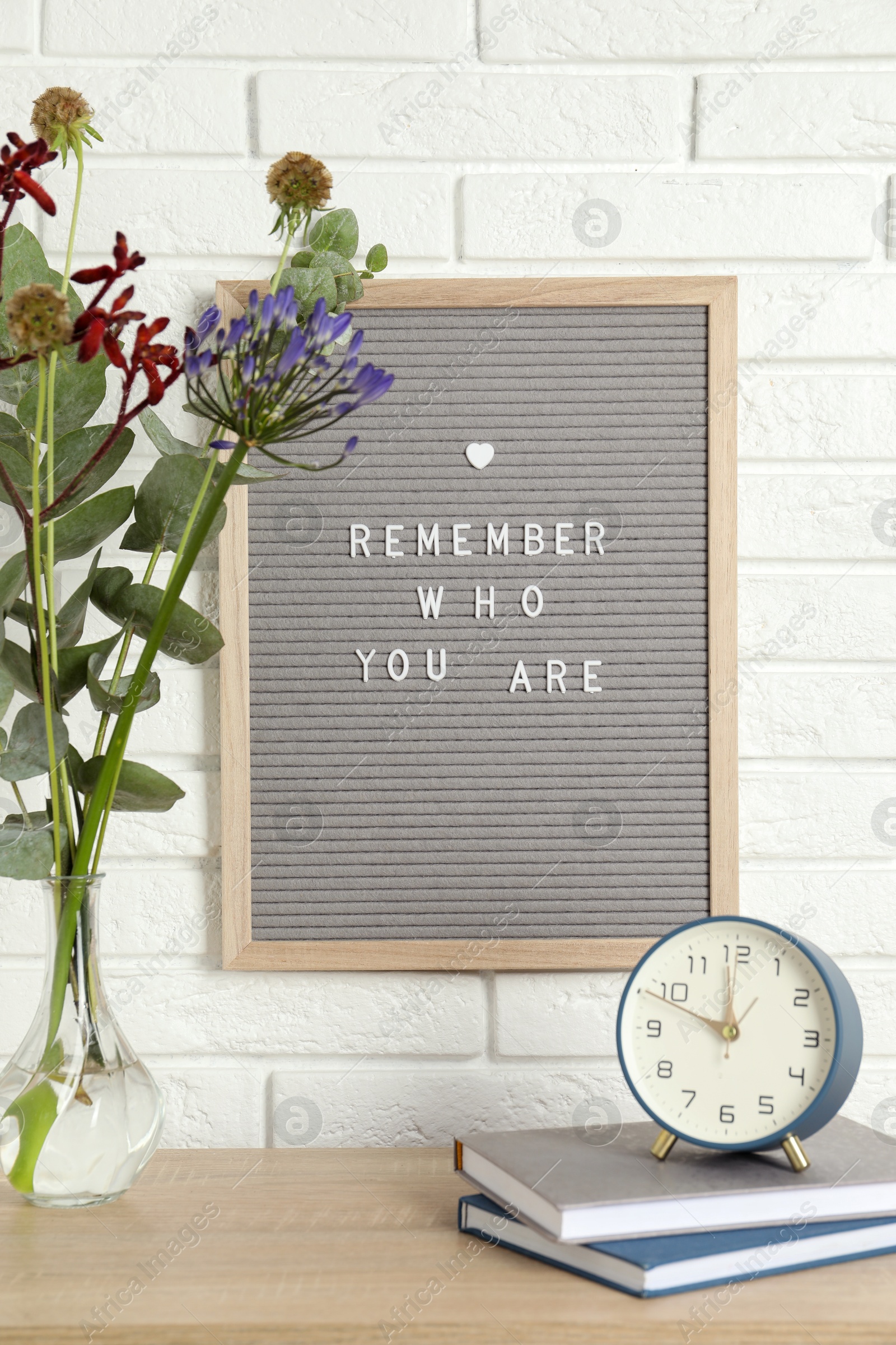 Photo of Letter board with phrase Remember Who You Are, books, alarm clock and bouquet in vase on wooden table indoors