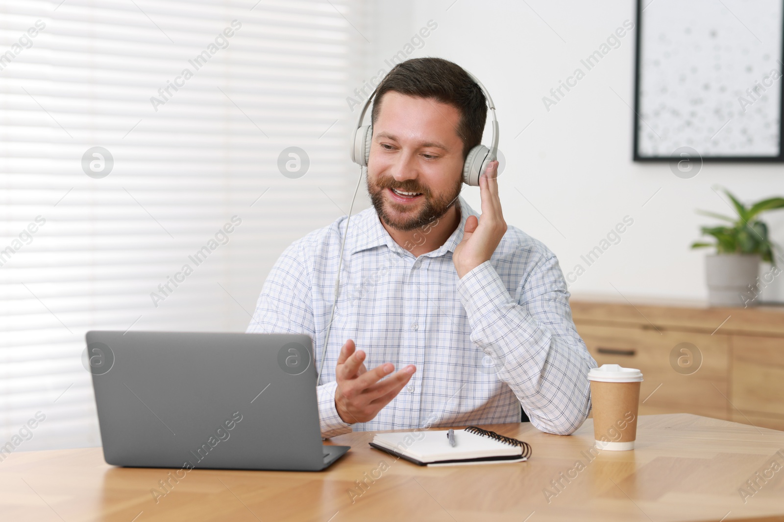 Photo of Interpreter in headphones having video chat via laptop at wooden table indoors