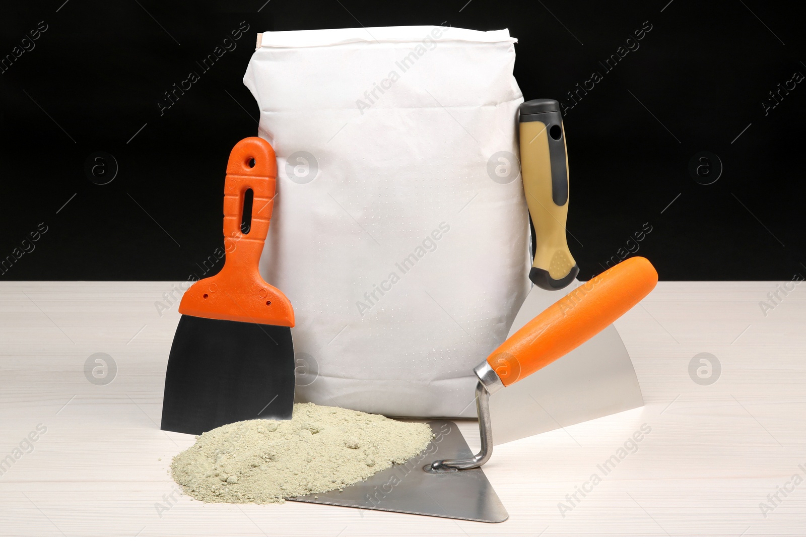 Photo of Bag of cement powder and putty knives on light wooden table against black background. Building material and construction tools