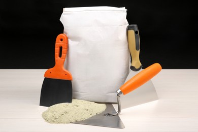 Photo of Bag of cement powder and putty knives on light wooden table against black background. Building material and construction tools