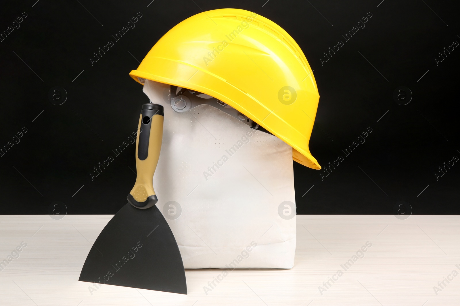 Photo of Yellow hard hat, bag of cement powder and putty knife on light wooden table against black background. Building material