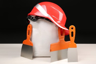 Photo of Red hard hat, bag of cement powder and putty knives on light wooden table against black background. Building material and construction tools