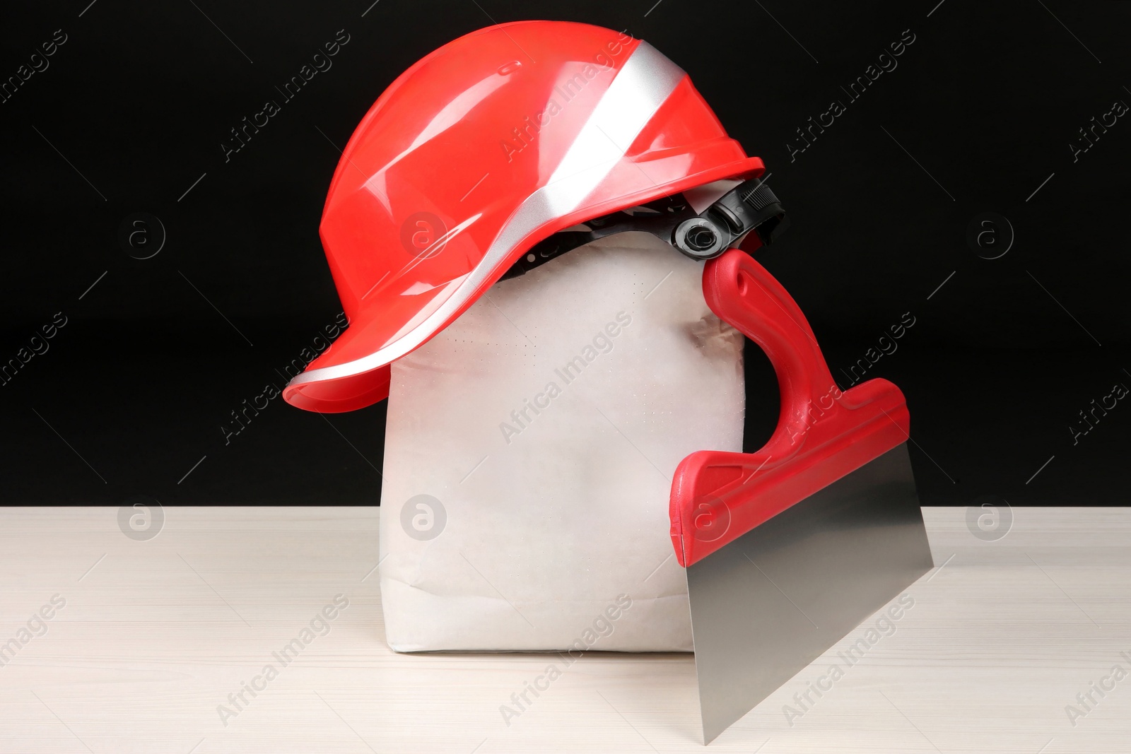 Photo of Red hard hat, bag of cement powder and putty knife on light wooden table against black background. Building material