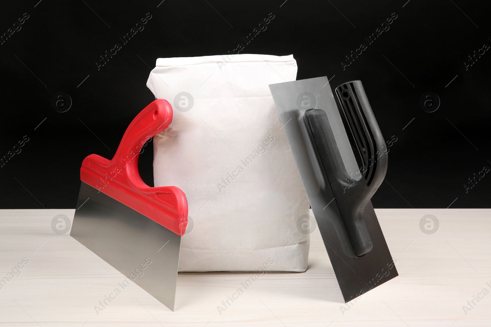 Photo of Bag of cement powder and putty knives on light wooden table against black background. Building material and construction tools