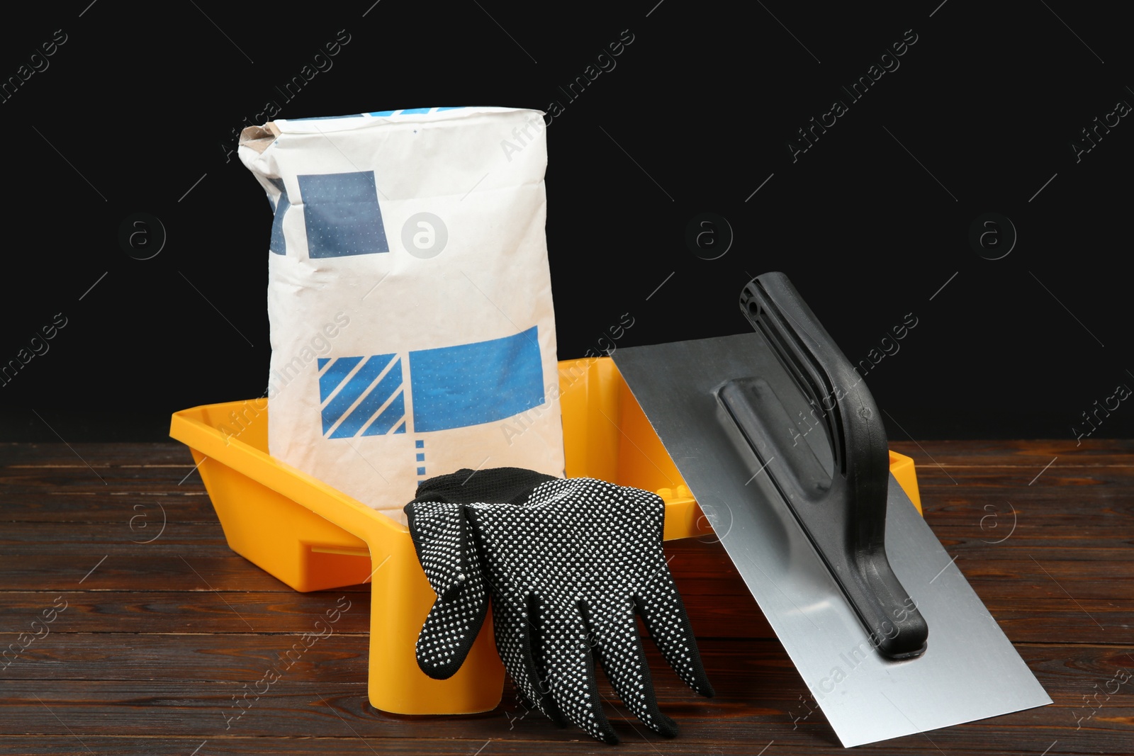 Photo of Bag of cement powder, tray, putty knife and rubber glove on wooden table. Building material and construction tools