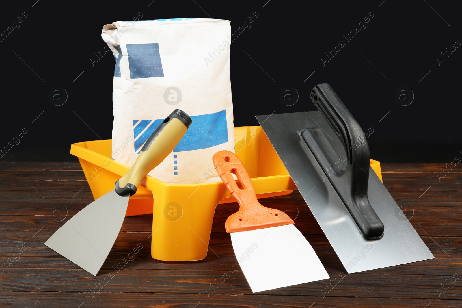 Photo of Bag of cement powder, tray and putty knives on wooden table. Building material and construction tools