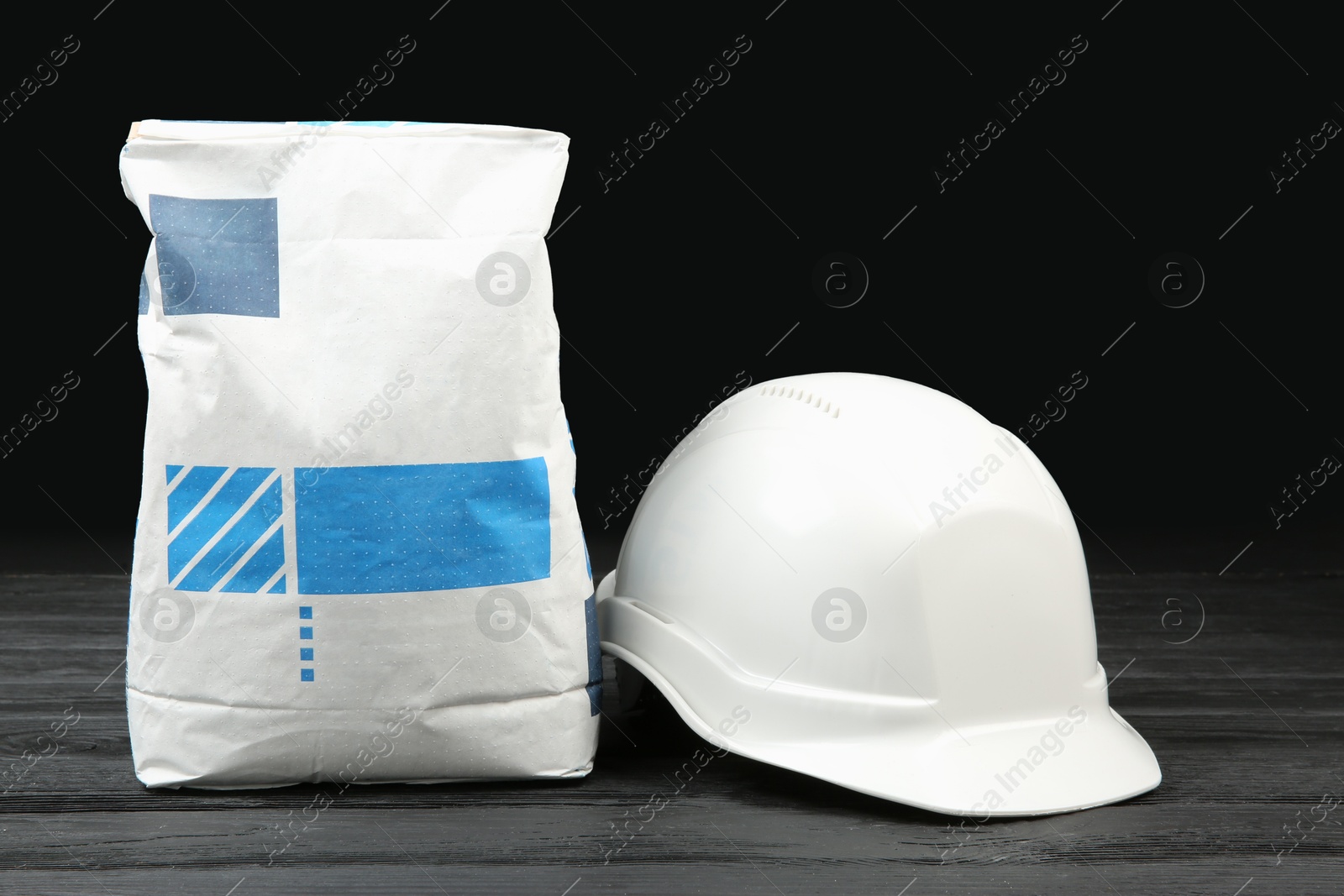 Photo of Bag of cement powder and white hard hat on dark wooden table. Building material