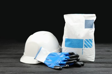 Bag of cement powder, white hard hat and rubber gloves on dark wooden table. Building material