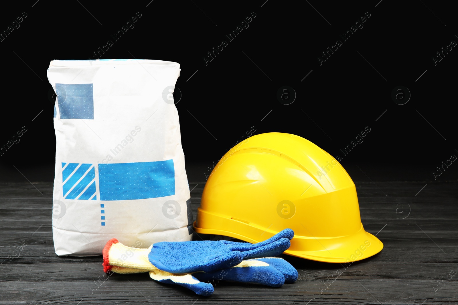 Photo of Bag of cement powder, yellow hard hat and rubber gloves on dark wooden table. Building material