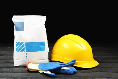 Bag of cement powder, yellow hard hat and rubber gloves on dark wooden table. Building material