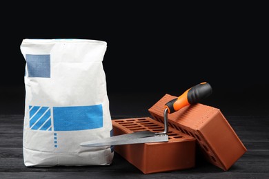 Photo of Red bricks, bag of cement powder and putty knife on dark wooden table. Building materials